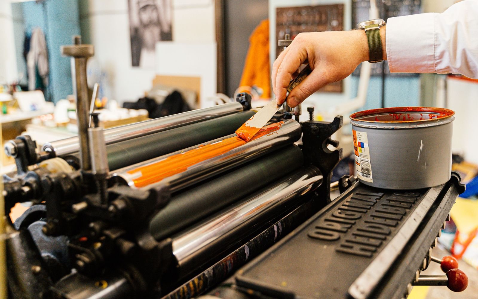 A hand spreading orange ink onto the rollers of a vintage printing press